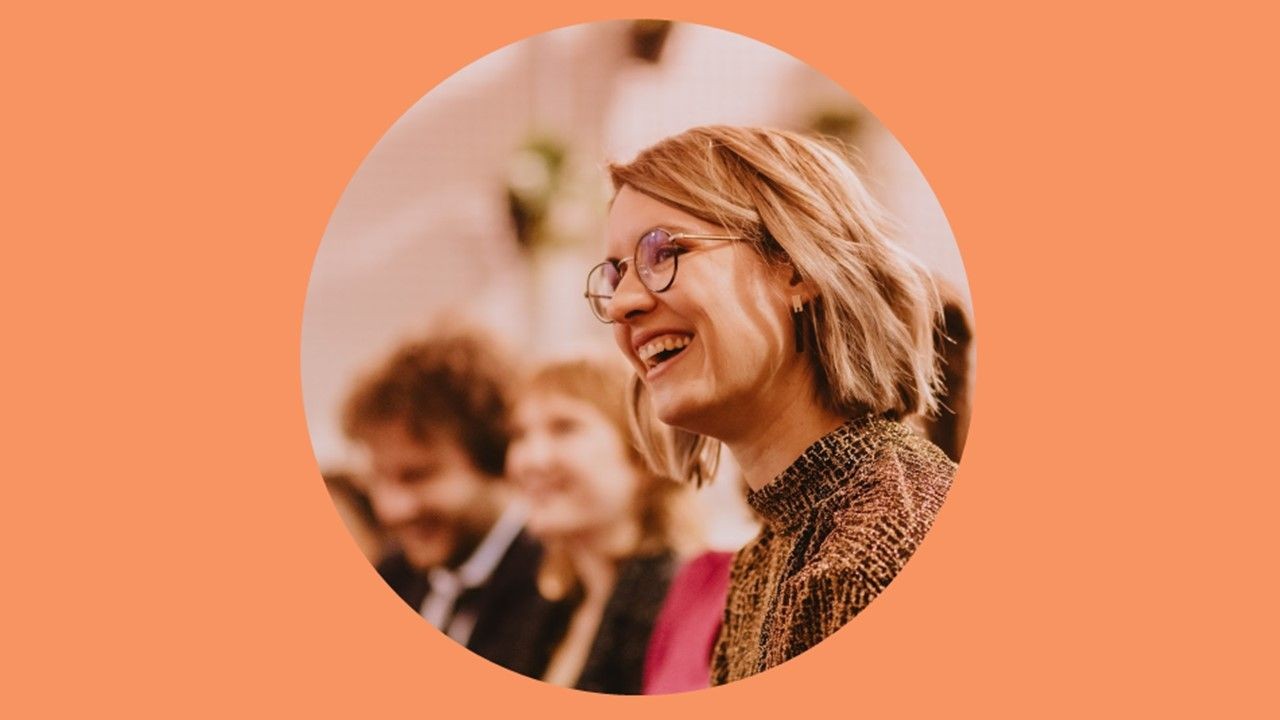 Smiling person with glasses in a crowded setting, against an orange background.